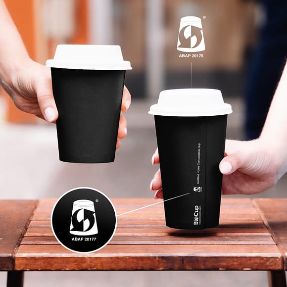Two hands holding up aqueous plastic-free BioCups. They’re black cups with white lids and white writing. The image zooms in on the compostable label, reading ABAP20177.