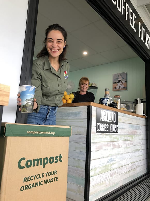 a woman working at a coffee shop. She’s holding a certified compostable coffee cup and placing it into the Compost Connect bin. She’s smiling at the camera.