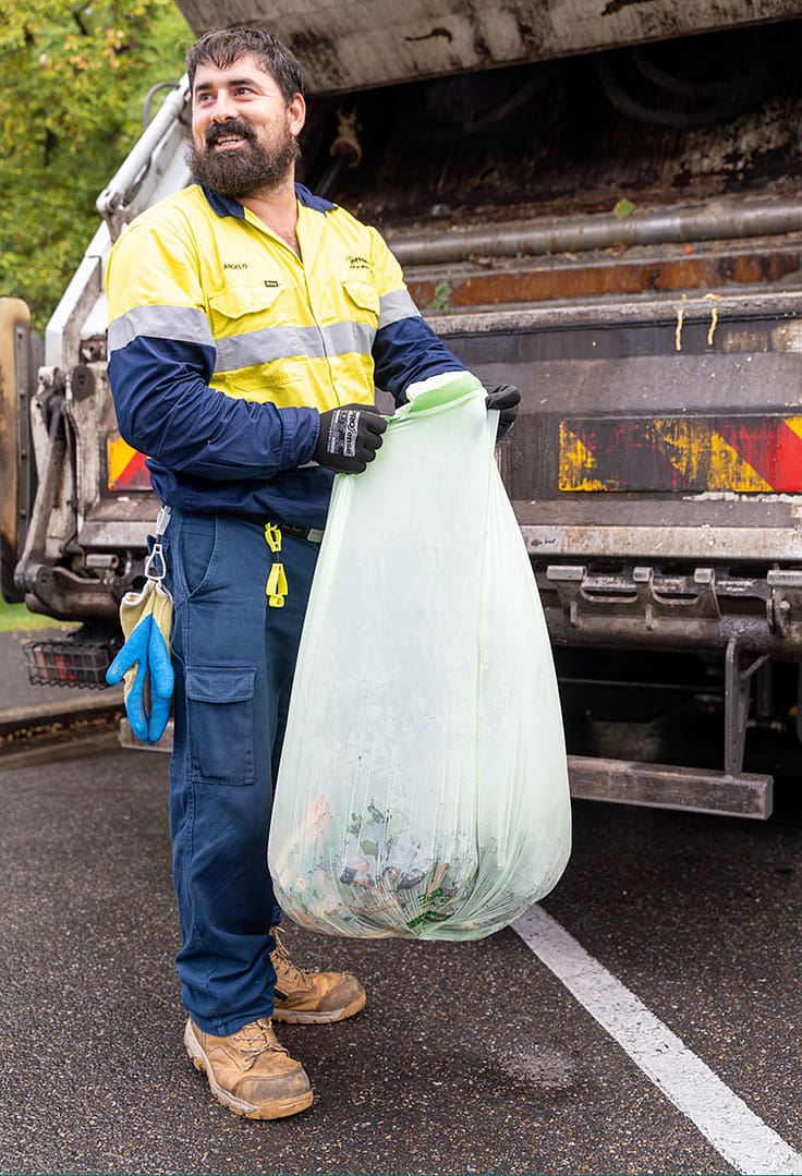 A worker behind a compost truck. He’s holding a compostable bag, wearing a high vis and smiling.